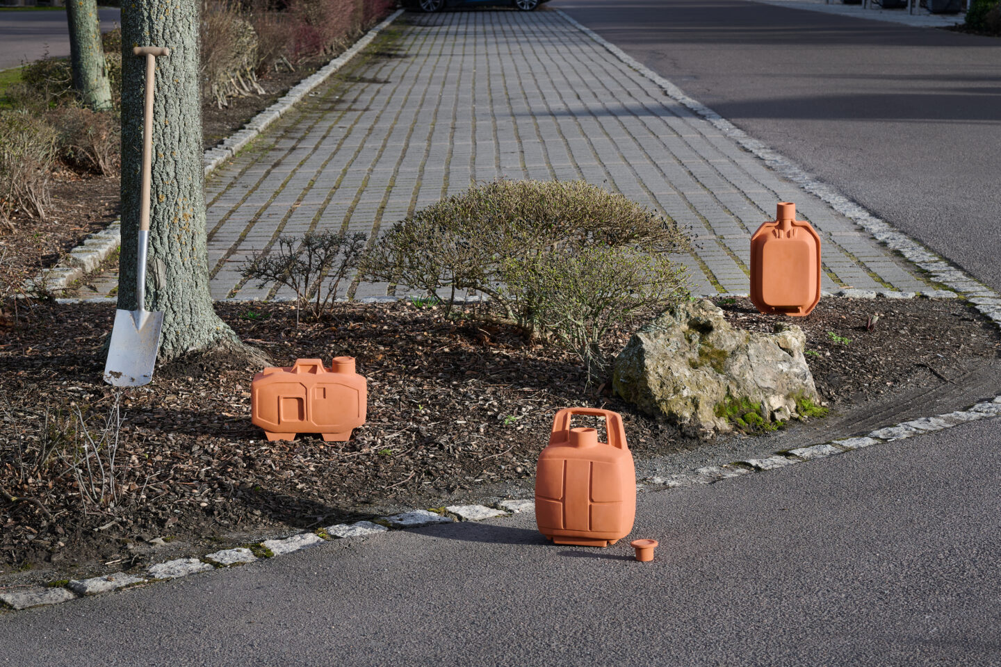 Terracotta containers arranged on a paved pathway, with a shovel leaning against a tree in the background.