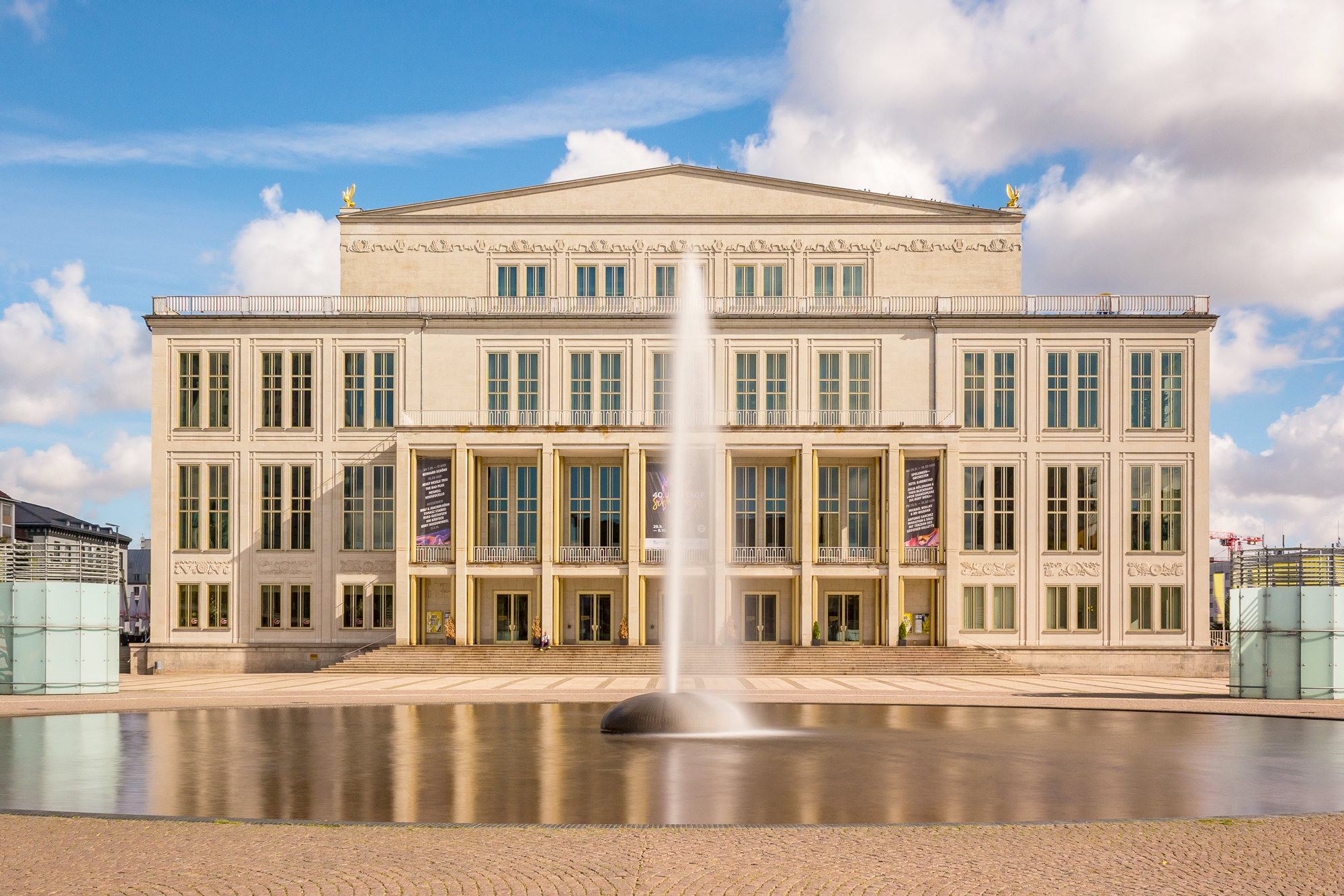 Opernhaus Leipzig with a central fountain, captured in long exposure, under a partly cloudy sky.