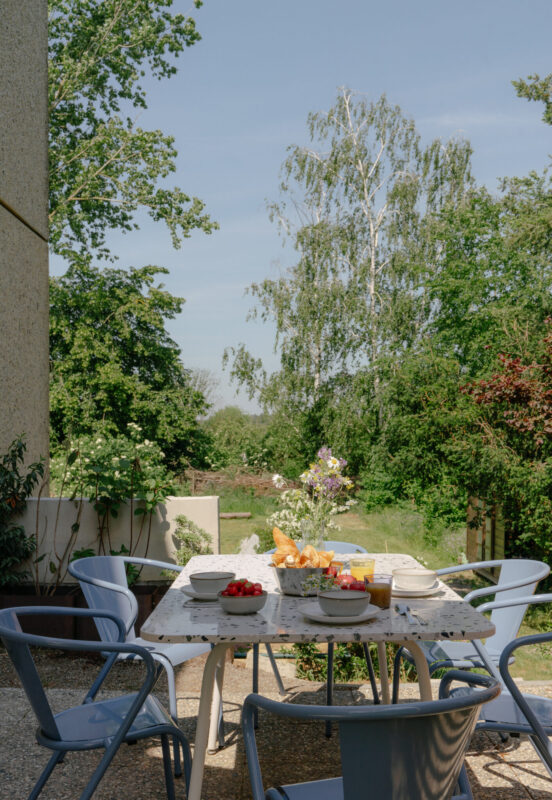 Table set with food and drinks outdoors, surrounded by green trees and plants, under a clear blue sky.