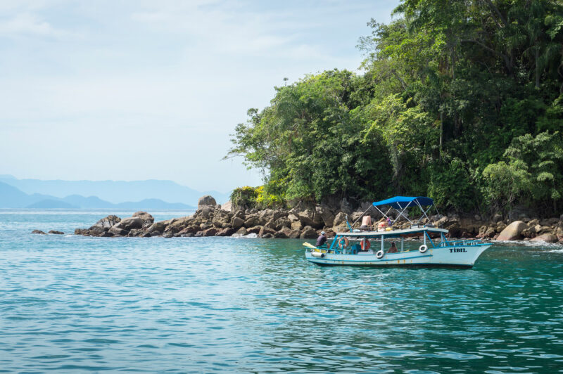 A small boat with a blue canopy floats near a rocky shore, surrounded by lush greenery and calm turquoise waters.