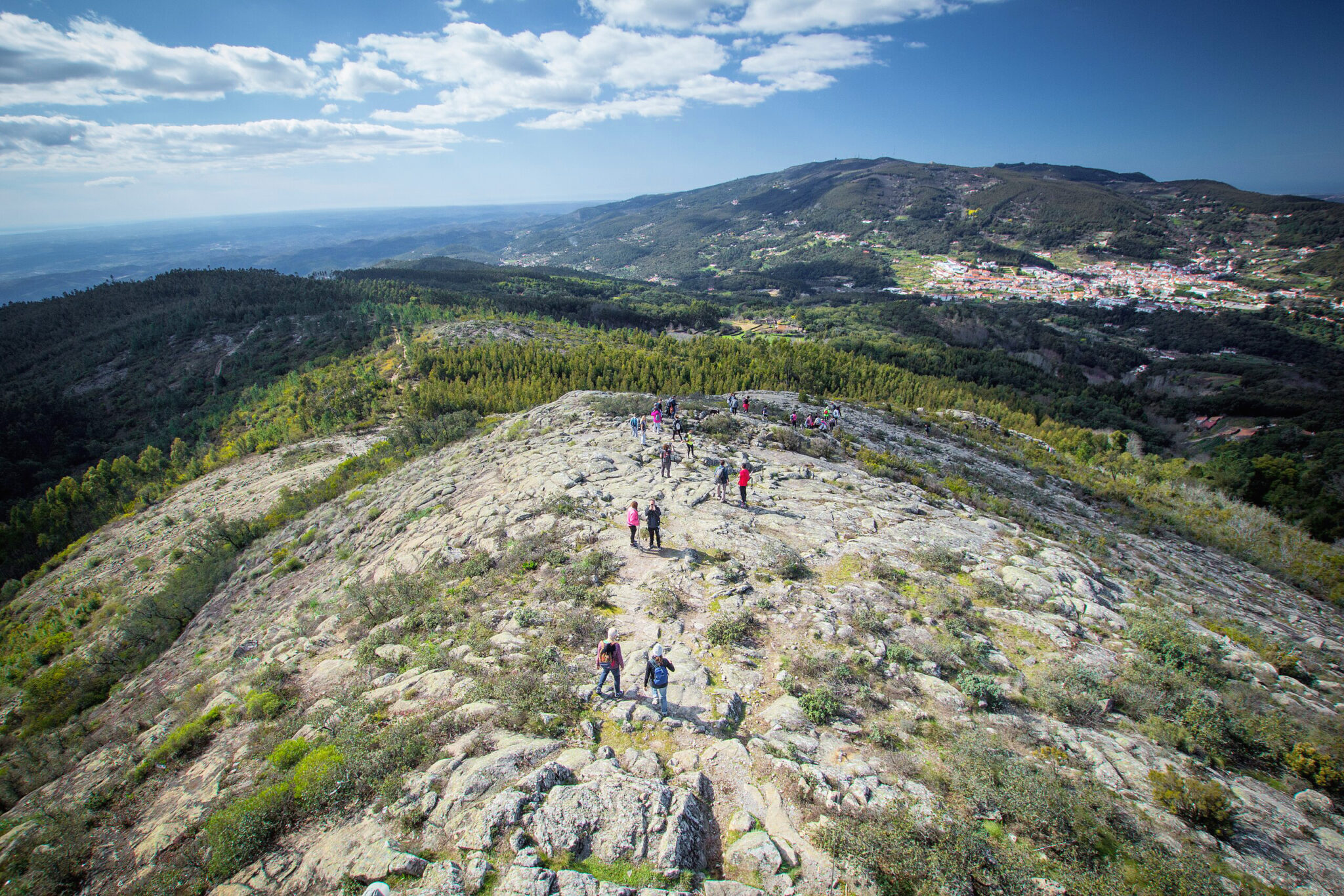 Hikers traverse a rocky hilltop with a panoramic view of green hills and a distant town under a blue sky.