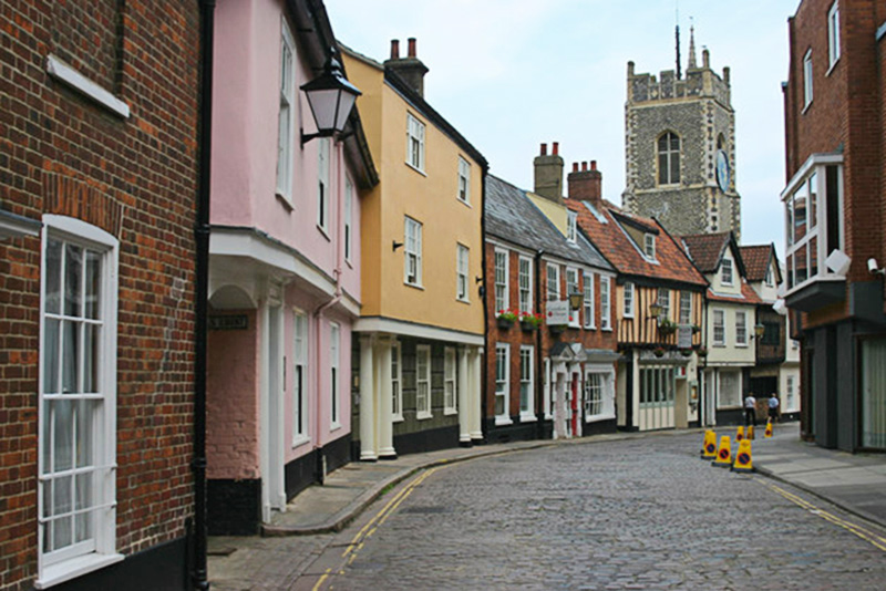 Cobblestone street in Norwich with colorful buildings and a church tower in the background.
