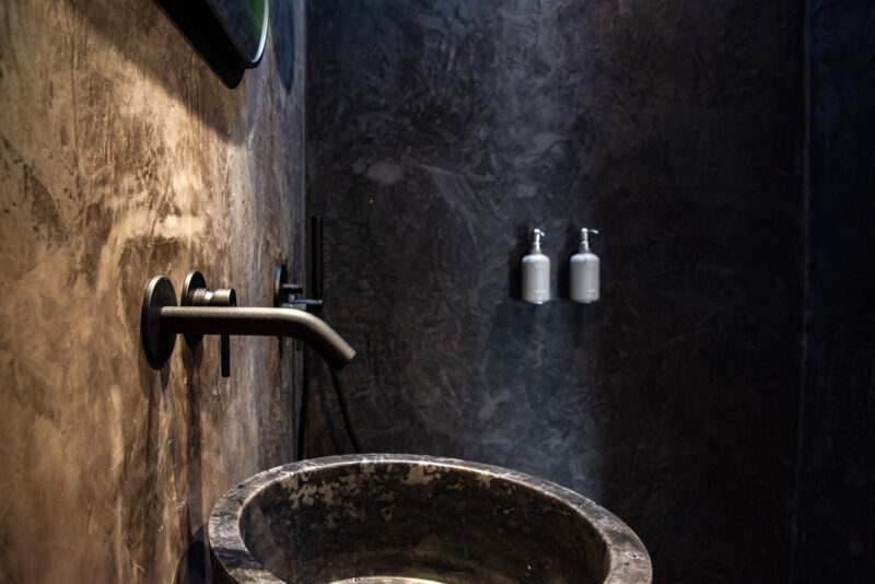 Modern bathroom with a stone basin, sleek black faucet, and two silver dispensers on a textured wall.