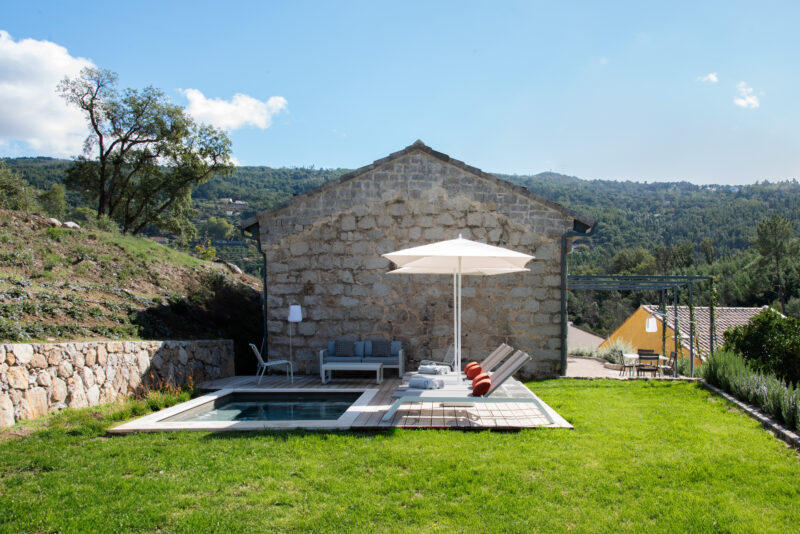 Modern stone house with a pool and lounge chairs, surrounded by greenery and hills under a clear blue sky.