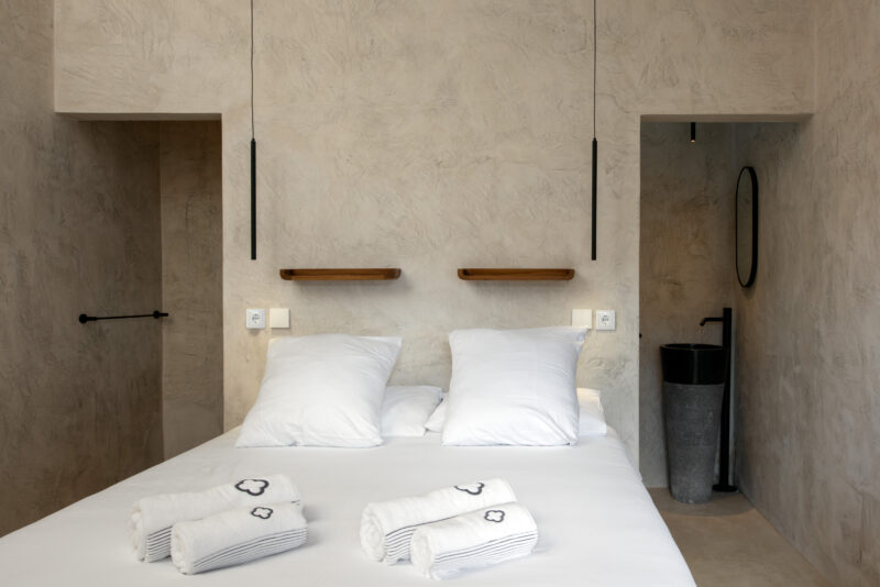 Minimalist bedroom with a double bed, two white pillows, and neatly rolled towels on the bed; features textured walls and ...