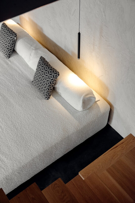 Cozy white bed with patterned pillows, softly lit, viewed from above, with wooden stairs in the foreground.