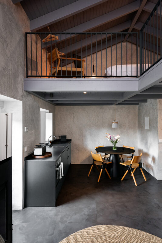 Modern interior of a loft-style space featuring a kitchen, dining area with a round table, and a lofted sleeping area.