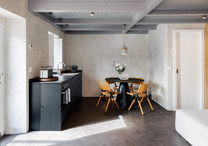 Modern kitchen with a black island, round table, and wooden chairs, featuring light walls and natural light.