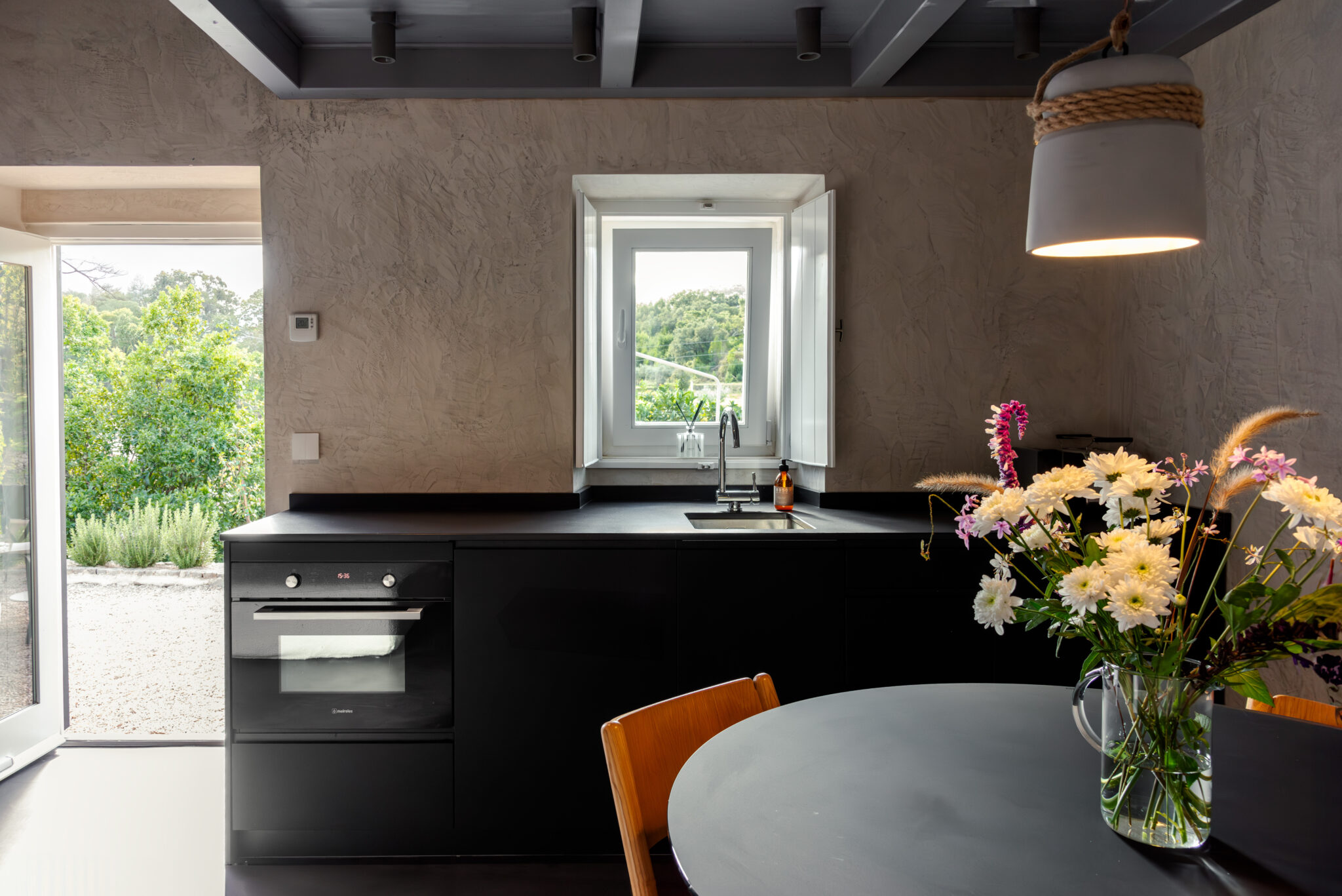 Modern kitchen featuring a black countertop, built-in oven, and a vase of flowers on a round table, with a window view of ...