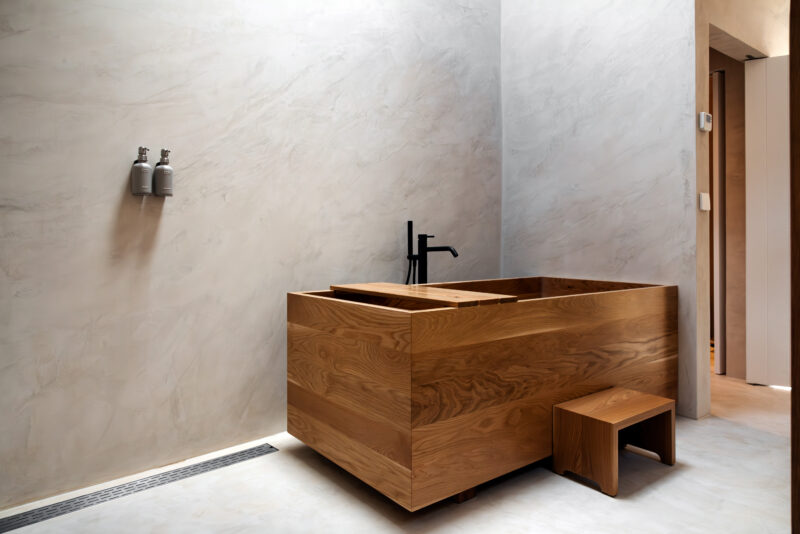 Wooden bathtub with a black faucet in a minimalist bathroom featuring smooth, light-colored walls.