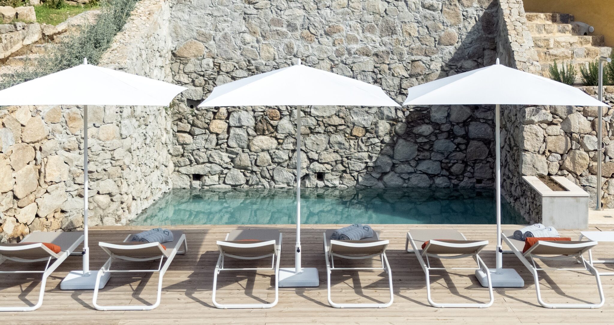 Lounge chairs with gray towels under white umbrellas beside a stone wall pool at The Yellow House.