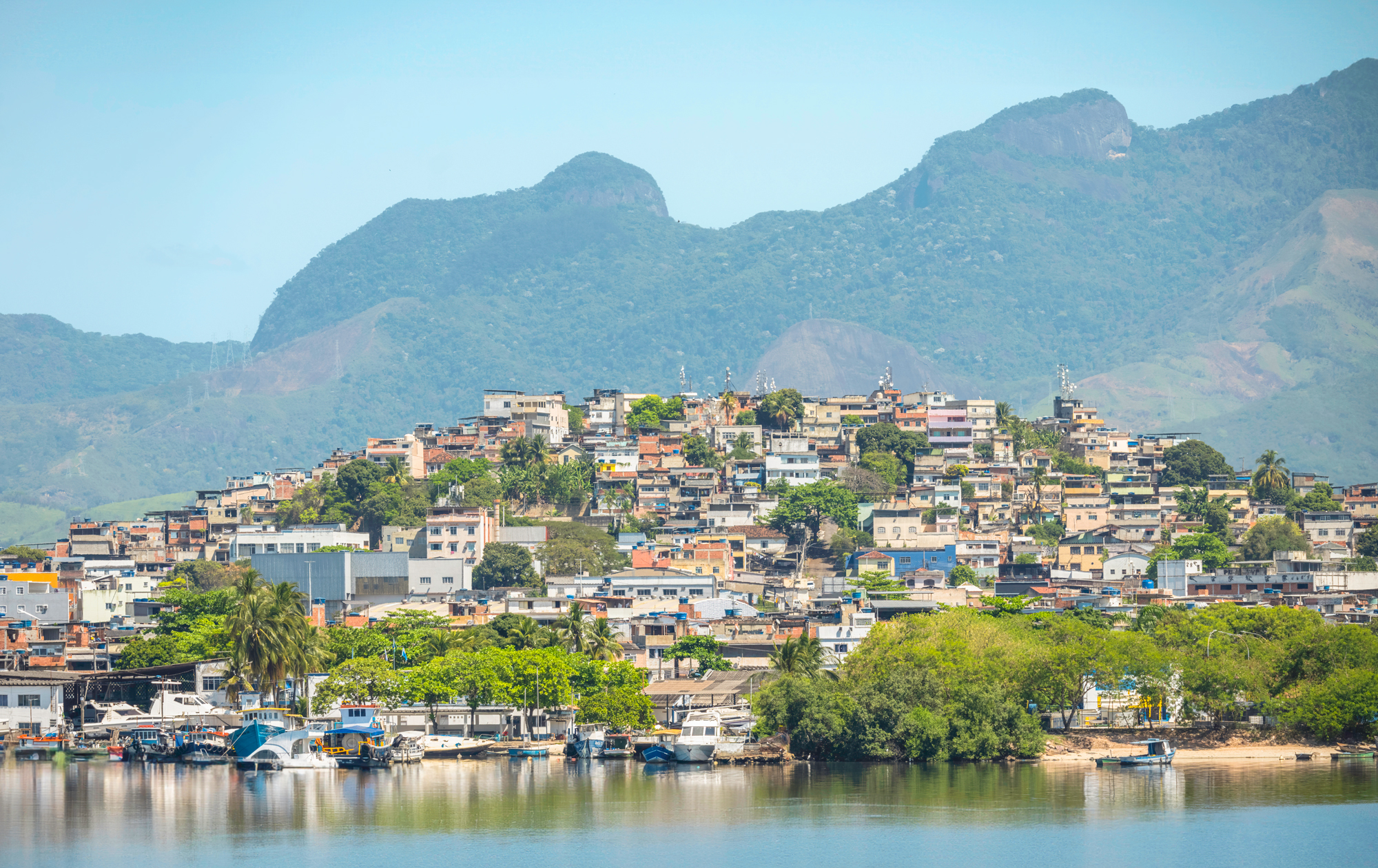 View of a Rio de Janeiro favela on a hillside, surrounded by lush greenery and mountains, with a calm water foreground.