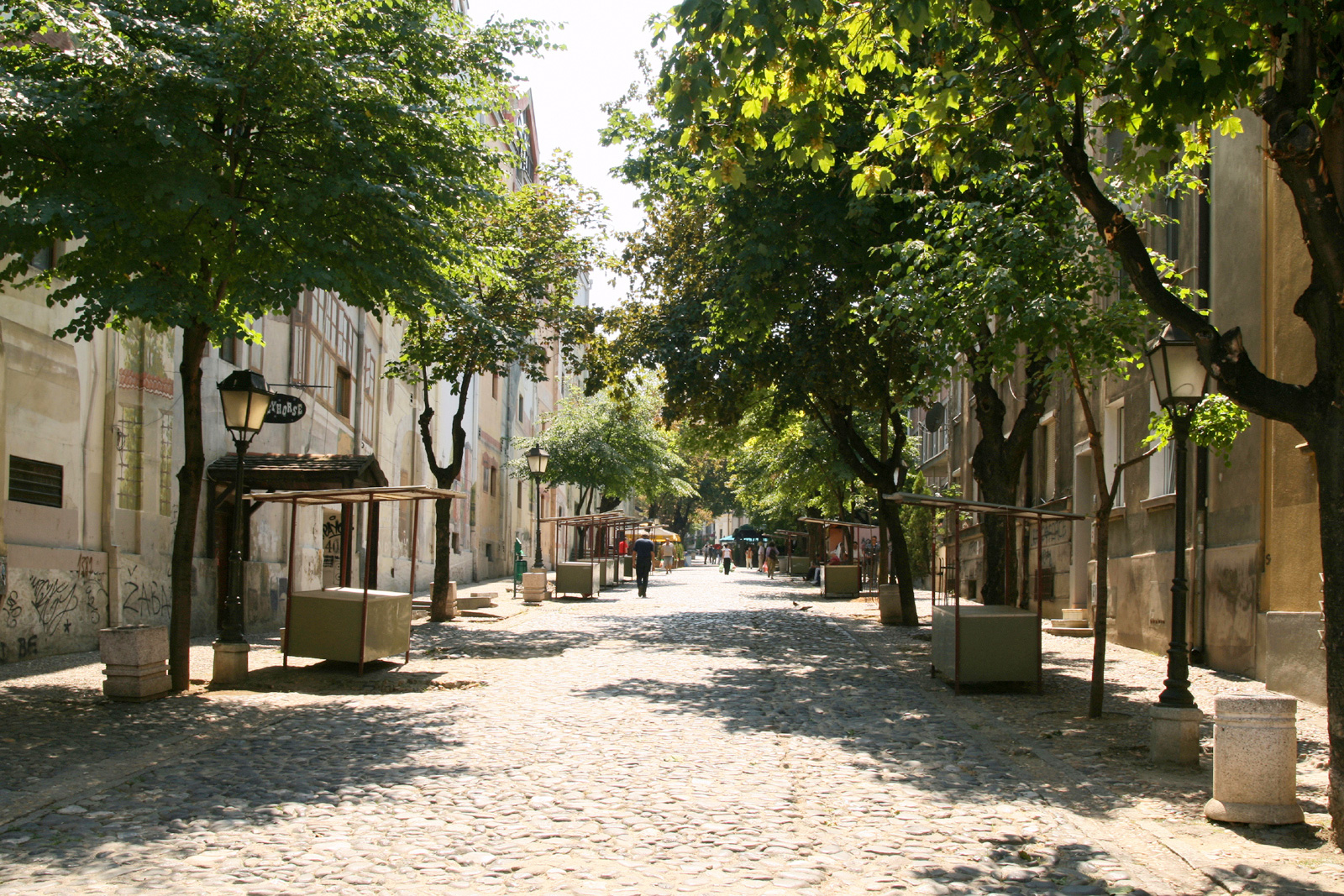 Cobblestone street in Skadarlija, Belgrade, lined with trees and empty market stalls under bright sunlight.