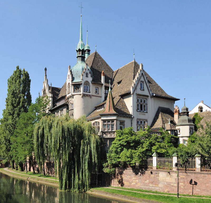 Historic building with a green spire, surrounded by trees and a canal, in Strasbourg.