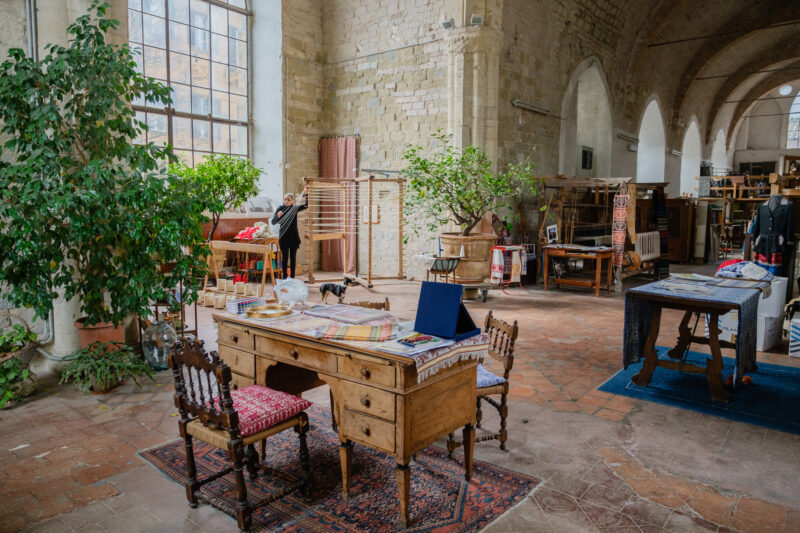 A spacious atelier featuring a wooden desk with colorful textiles, surrounded by plants, shelves, and various craft materi...