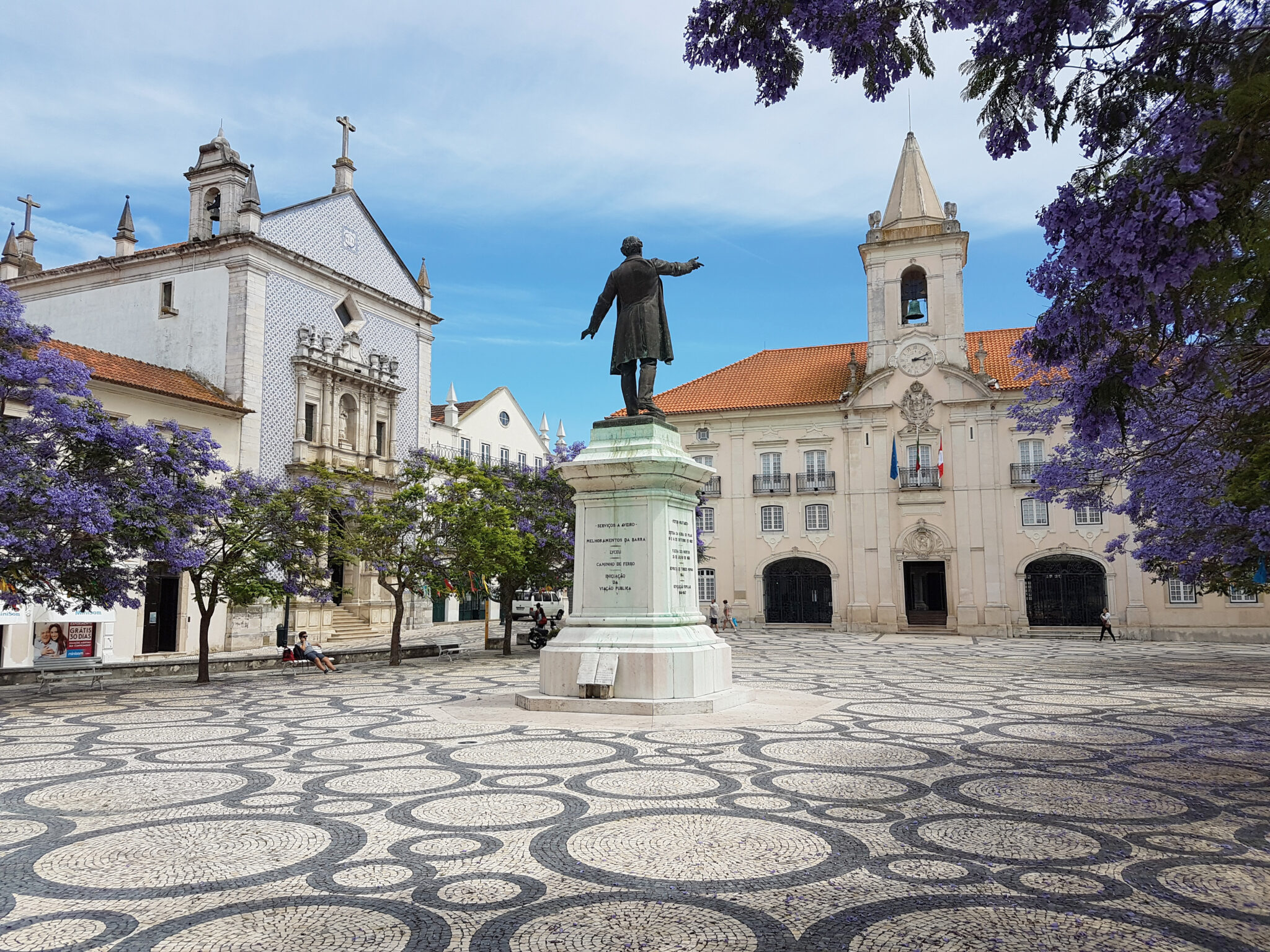 Statue in a town square surrounded by purple flowering trees and historic buildings under a blue sky.