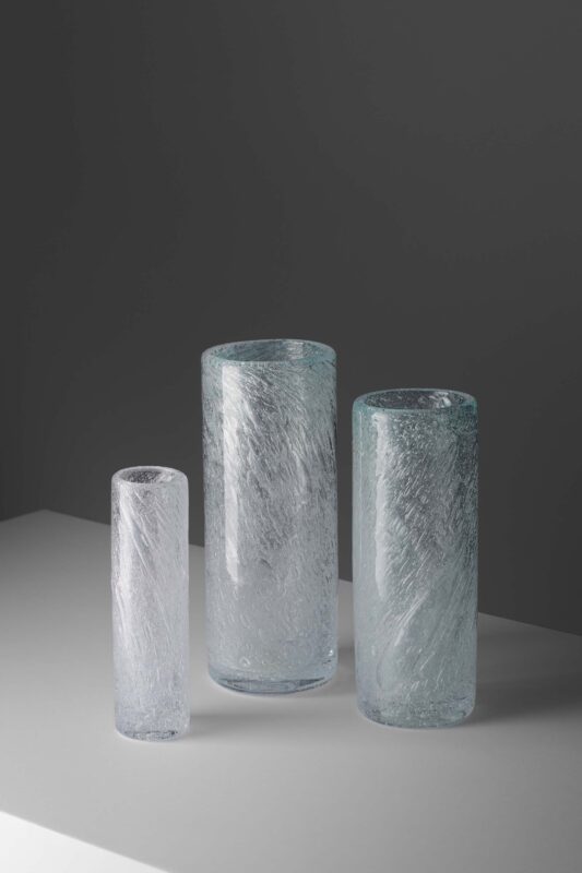 Three glass vases of varying heights, featuring textured surfaces, arranged on a light gray surface against a dark backgro...