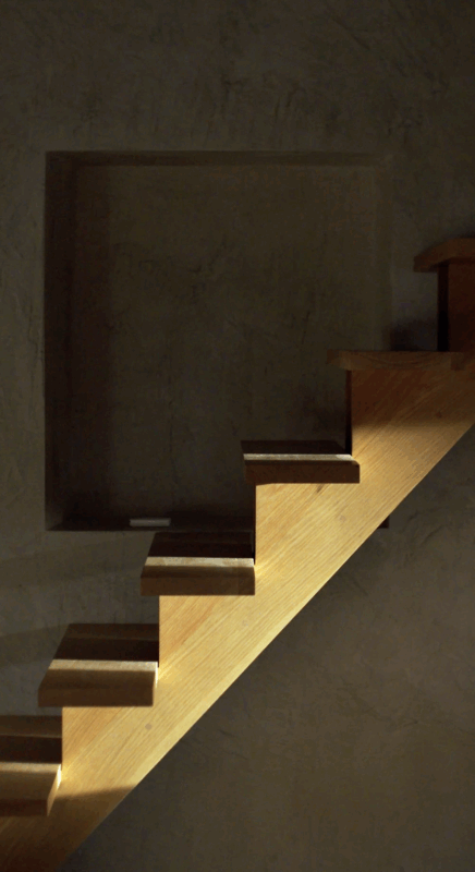 Wooden staircase with illuminated steps against a textured wall, featuring a recessed square niche above.