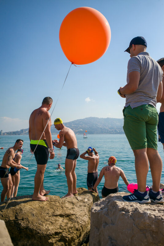 Swimmers prepare to enter the sea, some holding an orange balloon, with a coastal view in the background.