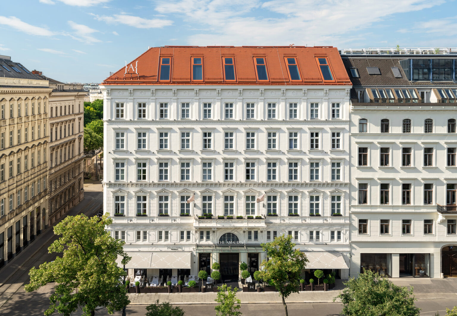 Elegant white facade of The Amauris hotel in Vienna, featuring a red roof and outdoor seating under trees.