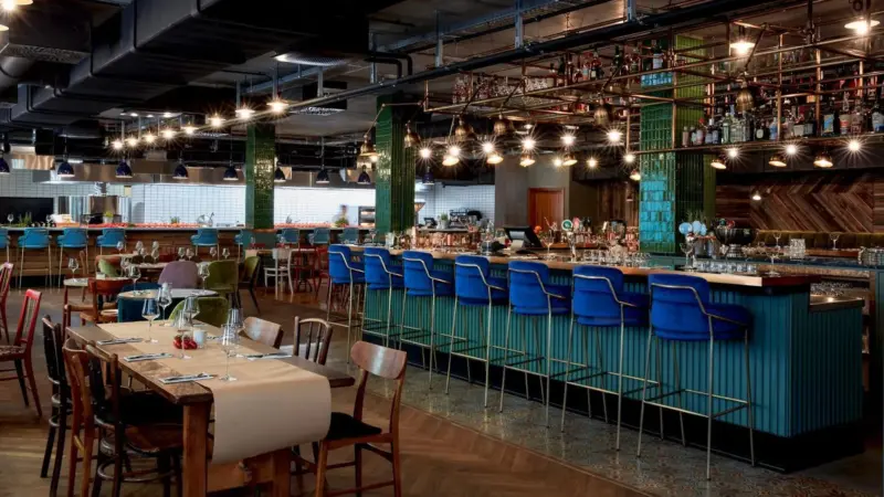 Stylish restaurant interior featuring a vibrant bar with blue stools, wooden tables, and decorative lighting.