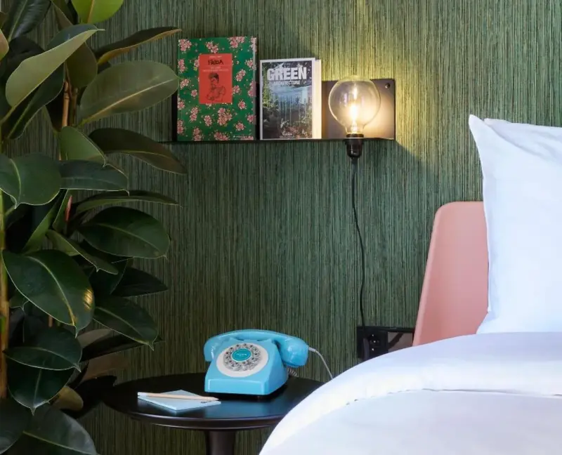A bedside shelf with two books, a light bulb, a vintage blue telephone, and a plant beside a bed with white linens.