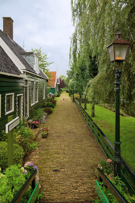 Pathway lined with greenery and traditional Dutch houses at Zaanse Schans, Netherlands, under a cloudy sky.