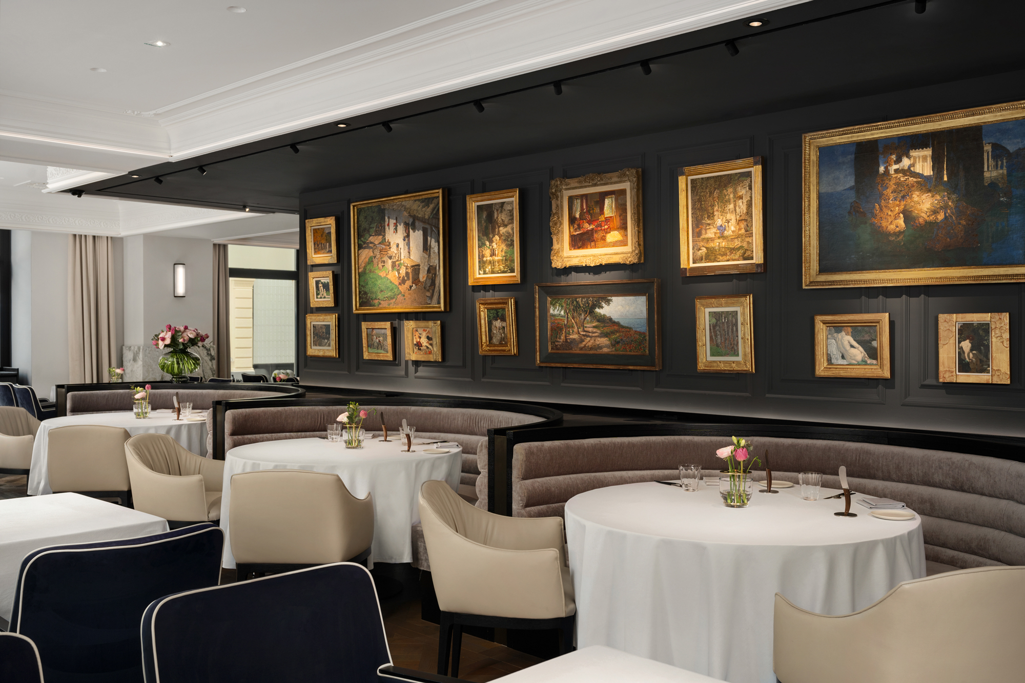 Elegant restaurant interior featuring round tables with white tablecloths, plush seating, and a gallery wall of framed art...