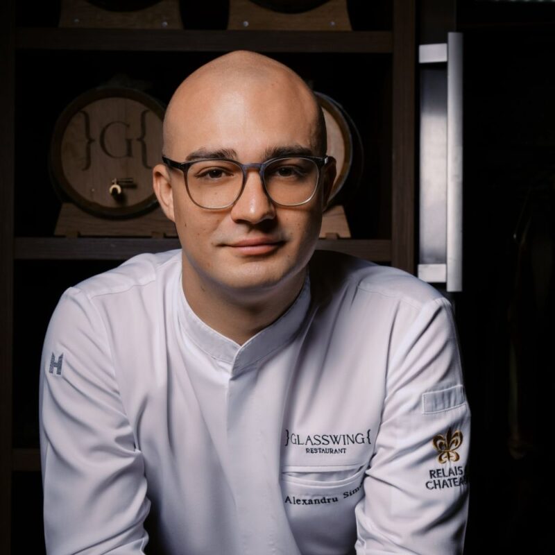 Chef Alexandra Simion in a white jacket, seated in front of a dark wooden backdrop with barrels.