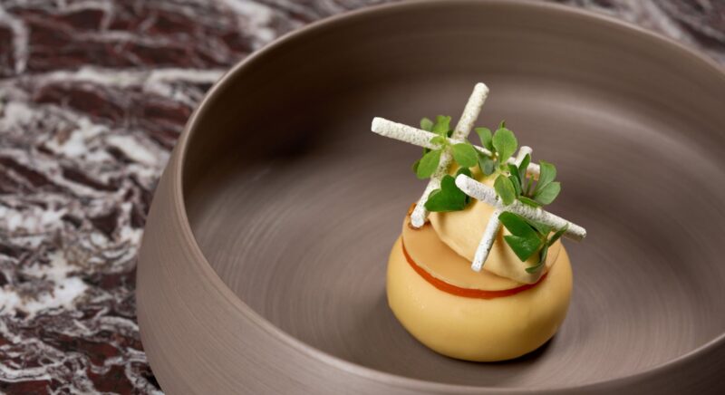 Dessert presentation featuring a round, yellow mousse topped with green herbs and white decorative sticks on a brown plate.