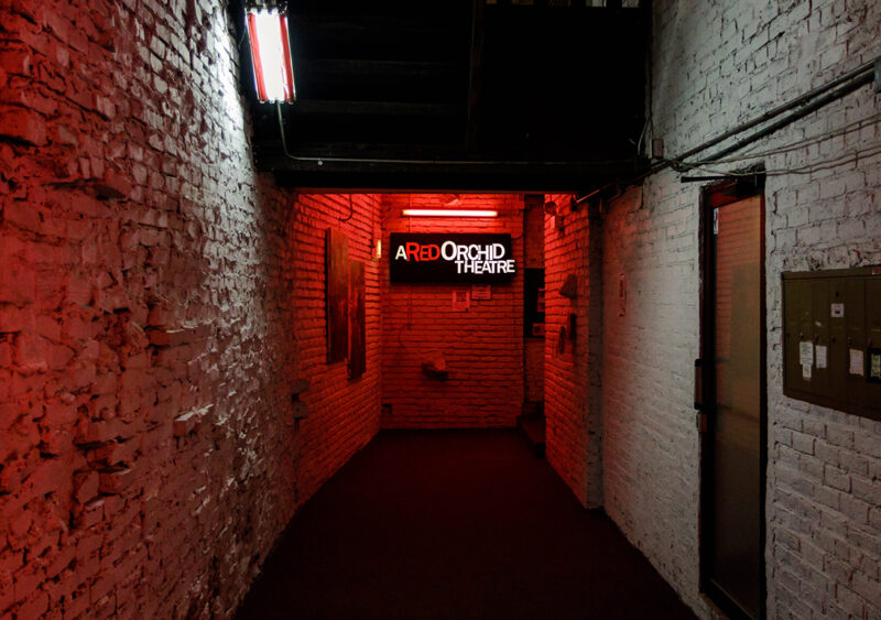 Dimly lit hallway with exposed brick walls, featuring a neon sign that reads "Red Orchid Theatre" in red light.