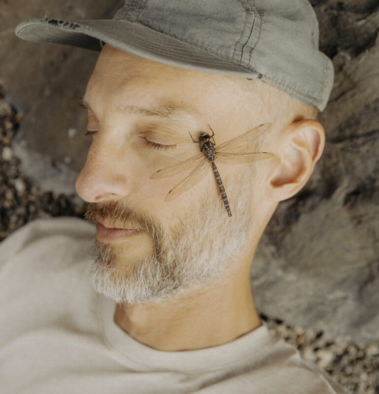 Man with a short beard and cap lying on a rocky surface, eyes closed, with a dragonfly resting on his cheek.