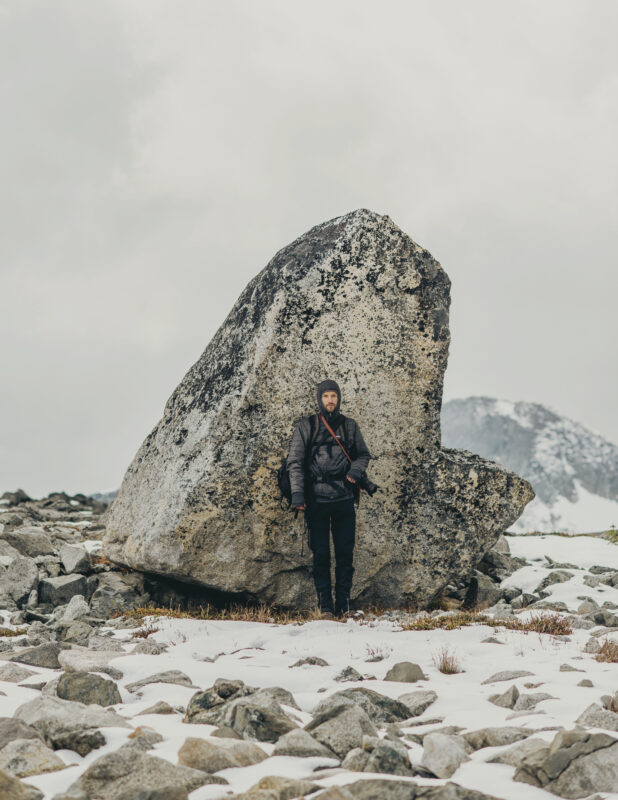 Man standing beside a large boulder in a rocky, snowy landscape with mountains in the background.