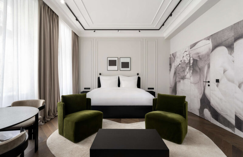 Modern hotel room featuring a large bed, two green armchairs, a round table, and a wall with artistic decor.