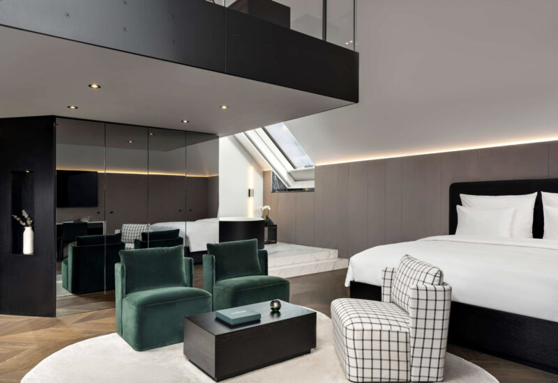 Modern hotel room featuring a bed, green armchairs, a coffee table, and mirrored walls, with a skylight above.