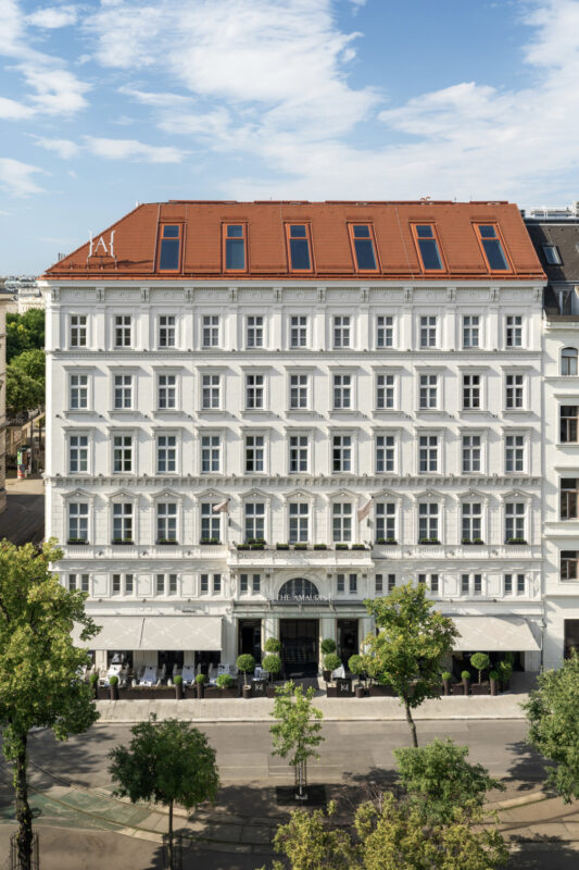 Elegant white hotel building with a red roof, featuring multiple windows and outdoor seating under canopies.