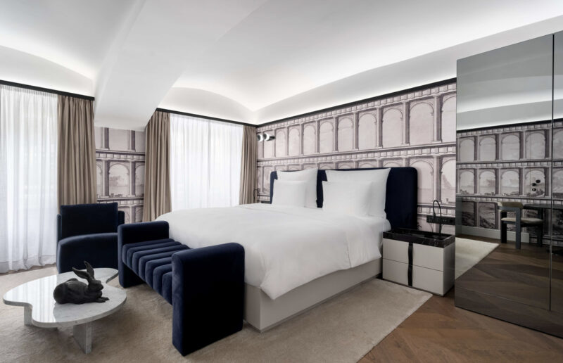 Modern hotel room featuring a white bed, navy blue seating, and a decorative wall mural with architectural details.