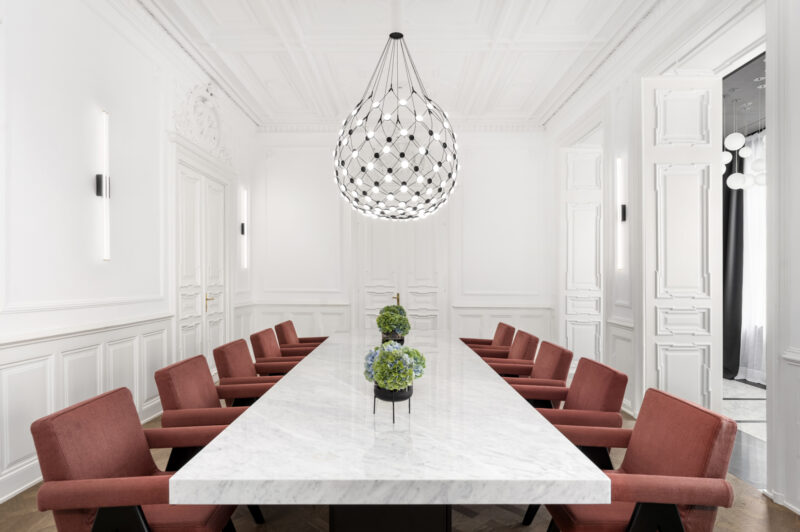 Modern conference room with a long marble table, red chairs, and a decorative pendant light, featuring greenery centerpieces.