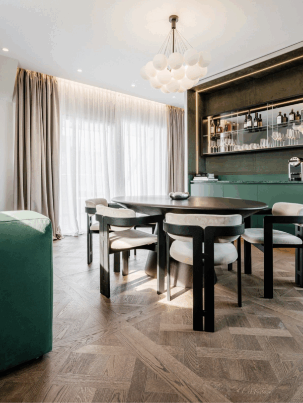 Modern dining area featuring a dark wooden table, stylish chairs, and a green accent wall with a bar. Natural light floods...
