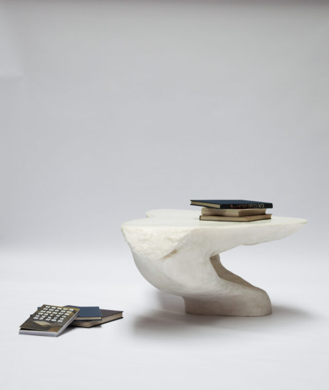 Sculptural white table with a curved base, topped with stacked books, against a plain background.