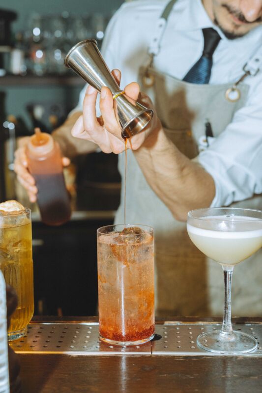Bartender pouring a dark syrup into a glass with a cocktail, another drink and a shaker visible in the background.