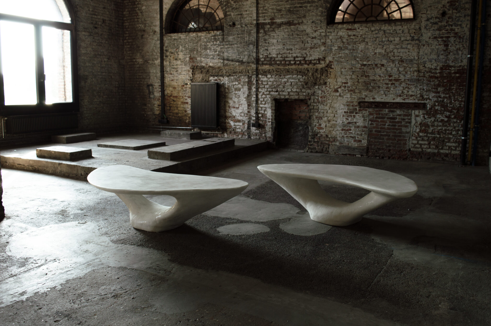 Two modern, sculptural white tables in an industrial space with exposed brick walls and large windows.
