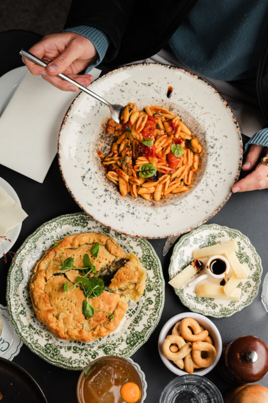 A hand holding a fork above a plate of pasta with vegetables, surrounded by a pie, cheese, and snacks on patterned plates.