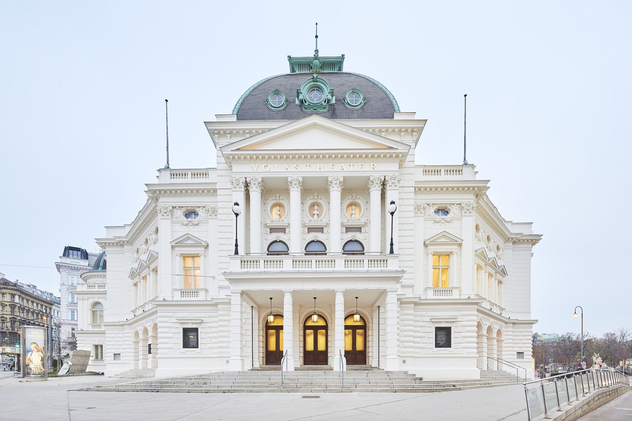 Historic Volkstheater building with ornate white façade and green dome, illuminated windows, against a pale sky.
