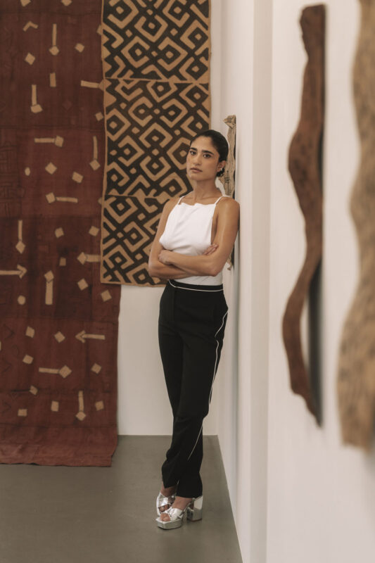 Woman in a white top and black pants stands against a gallery wall, with patterned artworks in the background.
