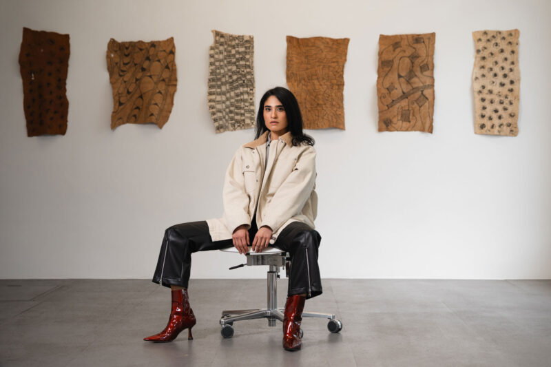 Woman in a beige jacket and red boots sitting on a stool in front of textured wall art.