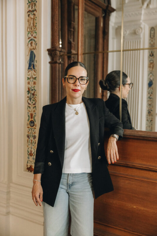 Stefanie Marik, general manager, poses in a stylish black blazer and white shirt, leaning against a wooden surface.