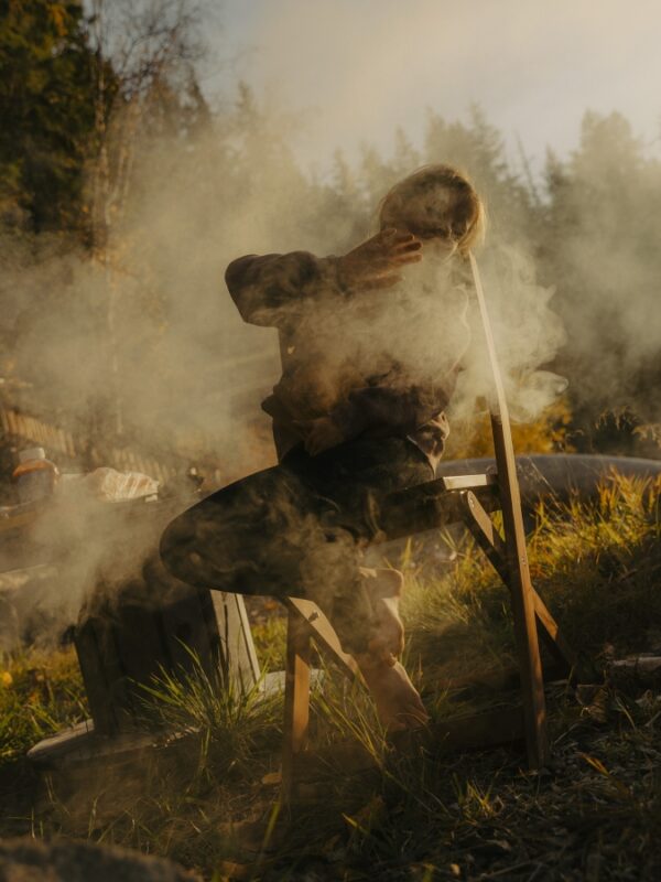 Person sitting on a wooden chair, partially obscured by smoke, surrounded by grass and trees in a natural setting.