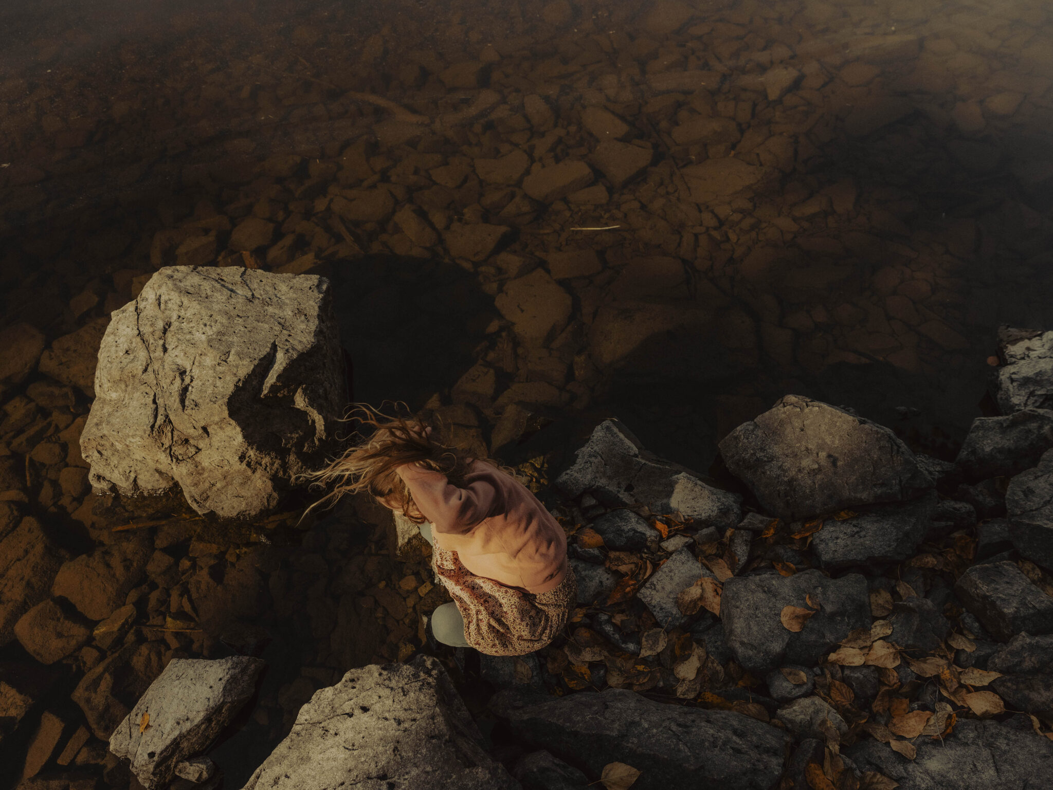 A person with long hair sits on rocky shore, surrounded by water and autumn leaves, in soft, natural lighting.