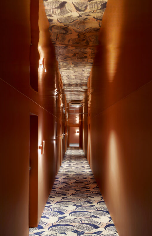 Long, narrow hallway with a patterned carpet and warm orange walls, illuminated by wall sconces.